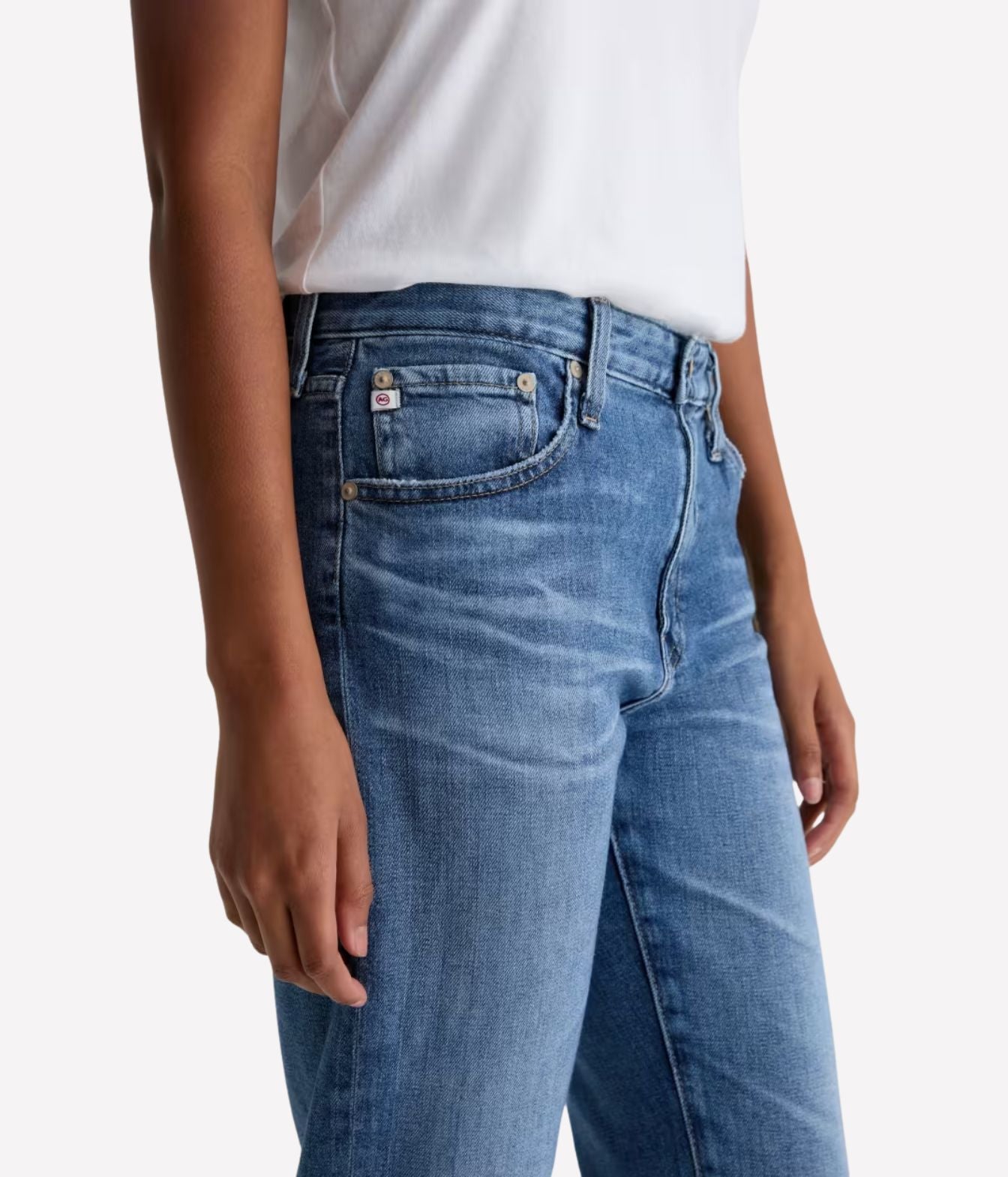 Close-up of a person wearing blue denim jeans with a white top, shown from the waist down against a light gray studio background.