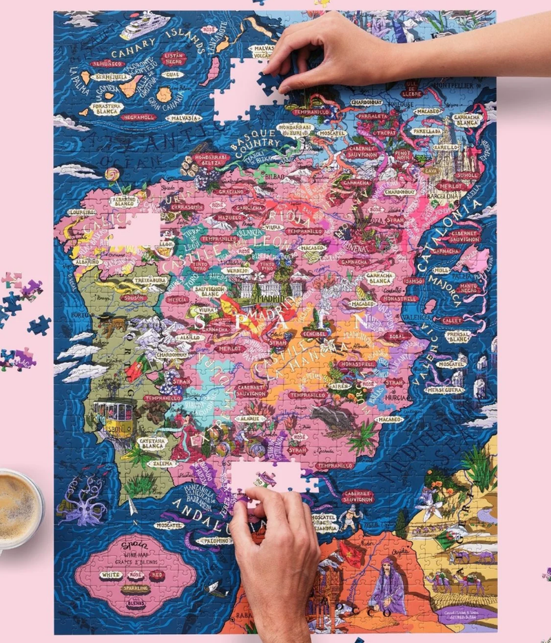Hands placing the last pieces into the Water and Wines 1000 Piece Puzzle of Spain, showing the hand-illustrated map of wine regions and grape varieties beside a cup of coffee