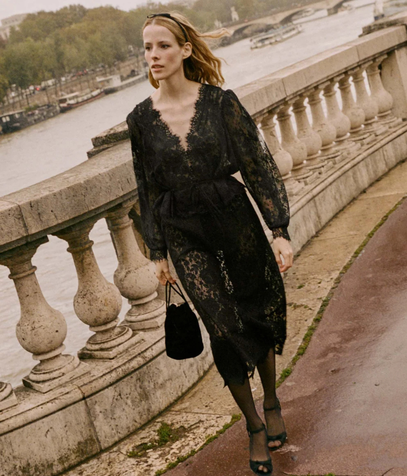Model walking in the DÔEN Amaline Top In Black with a matching black lace midi skirt, sheer black tights, black strappy heeled sandals, a black velvet pouch bag and a black headband