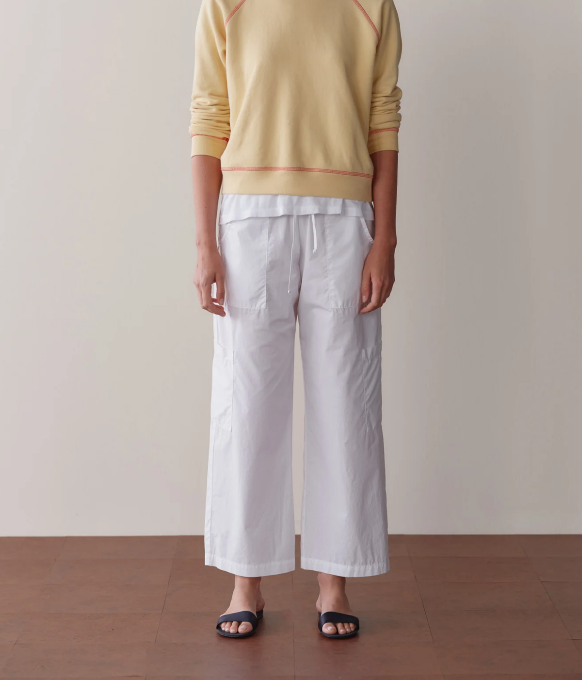Front view of a model wearing the DONNI. The Pop Drawstring Cargo Pant in Powder with a pale yellow raglan sweatshirt with orange contrast stitching over a white tee and navy slide sandals