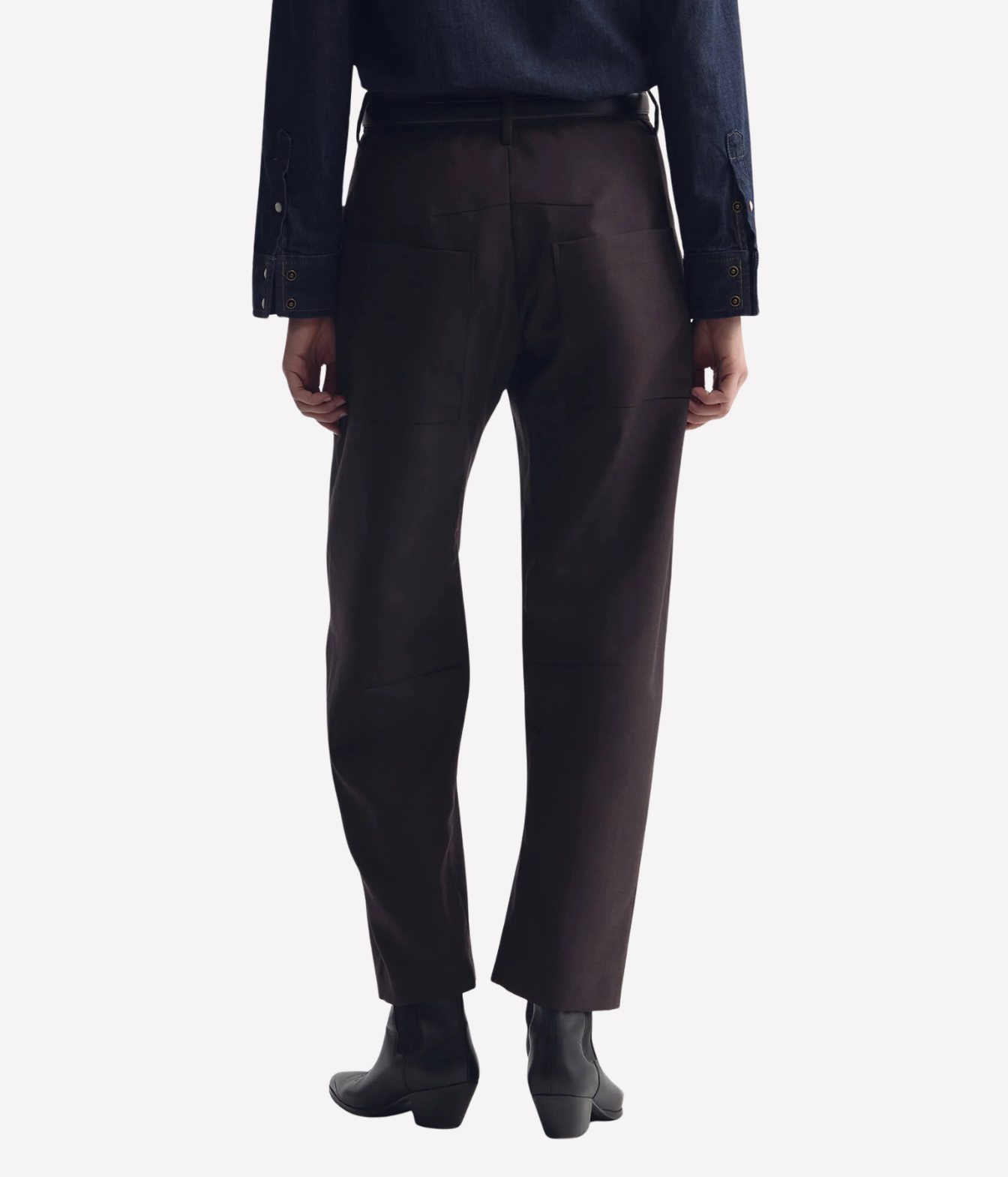 Rear view of a person wearing dark brown straight-leg trousers, a dark blue shirt, and black ankle boots against a plain white background.