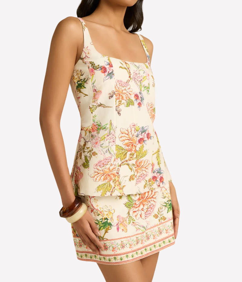 A model wearing a sleeveless floral print mini dress with thin straps and a tiered peplum waist, shown against a light gray studio background.