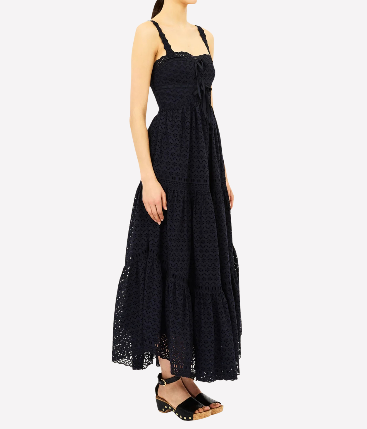 A woman modeling a black lace maxi dress with thin shoulder straps, a tie at the bust, and a tiered skirt, paired with black heeled sandals against a light gray background.