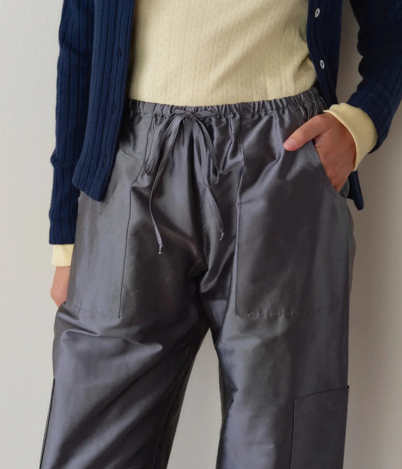 Detail of the DONNI. The Silk Taffeta Cargo Pant in Pewter, showing the sheen of the silk taffeta, the gathered drawstring waist and stitched front pockets, worn with a pale yellow pointelle top and navy cardigan