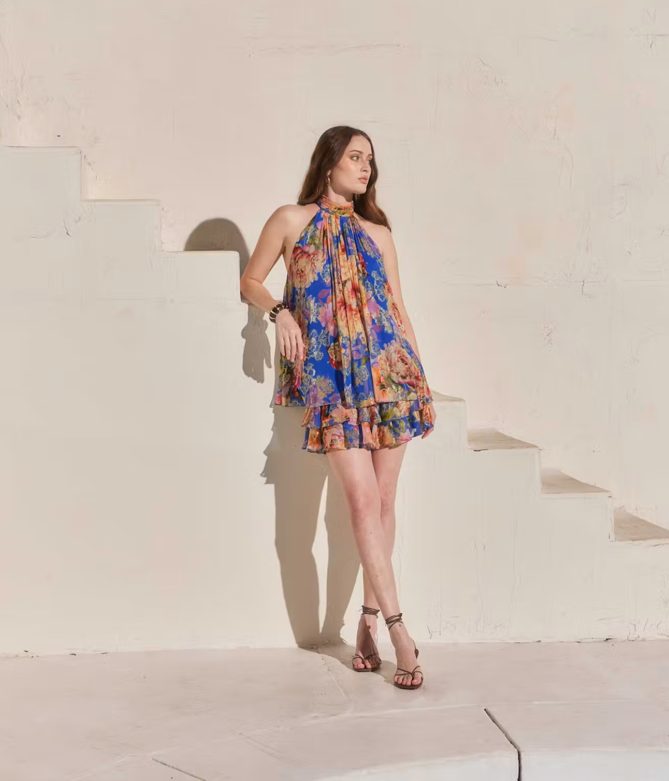 A woman models a colorful floral halter mini dress with a ruffled hem, standing on white concrete stairs against a textured beige wall.