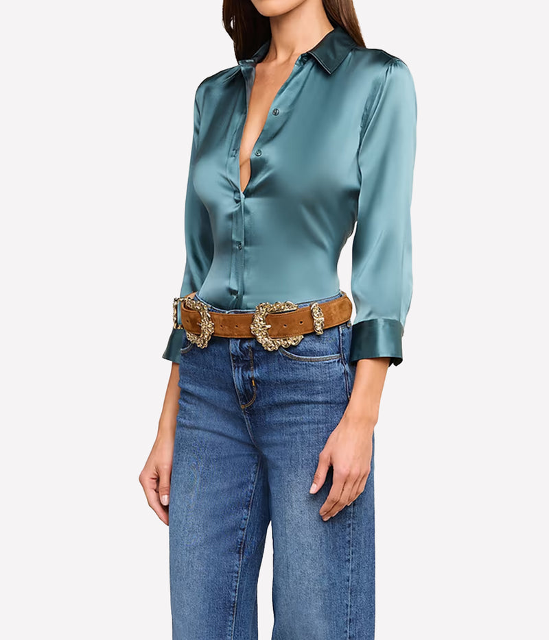 A woman wearing a cadet blue silk button-up blouse with three-quarter sleeves, tucked into blue jeans with a brown western-style belt featuring large ornate gold-tone buckles.