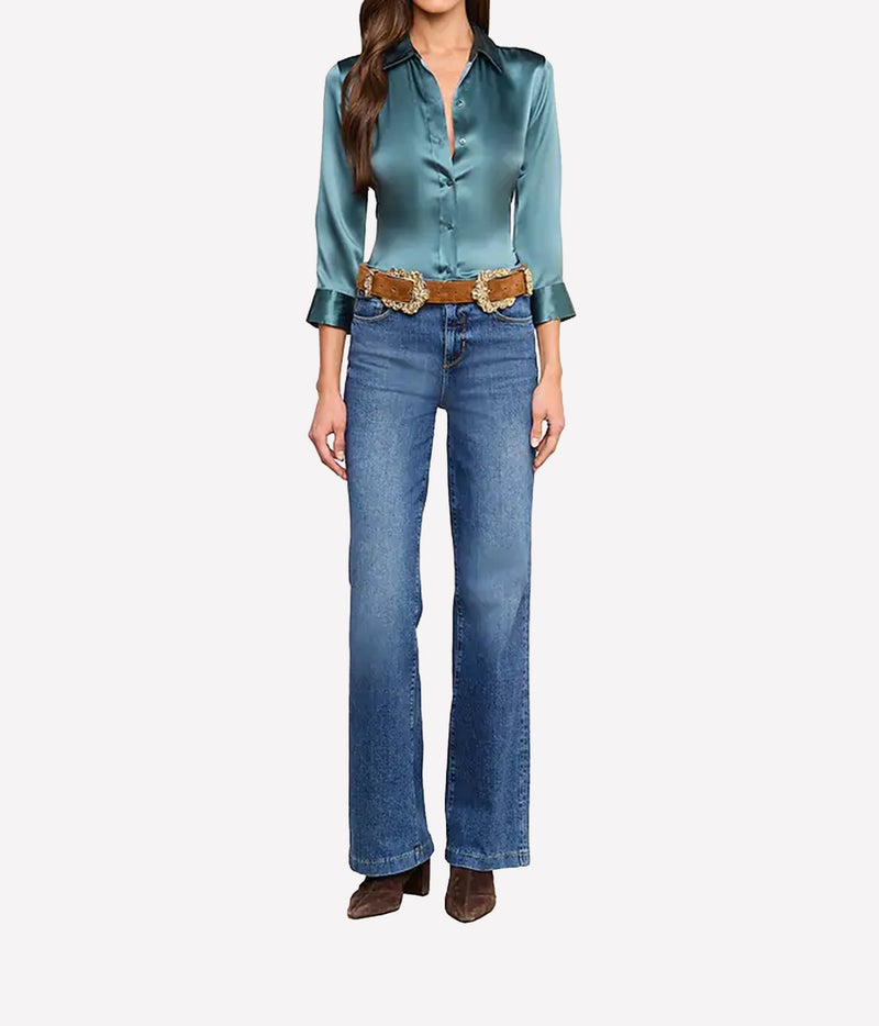A model wearing a cadet blue silk button-down shirt, blue flare jeans, a brown belt with gold hardware, and brown boots.