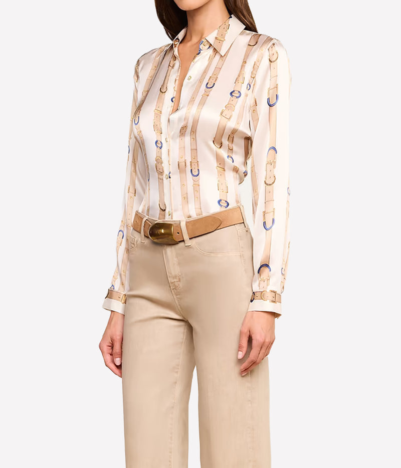 A woman wearing a cream silk button-up shirt with a gold and blue leather strap print, paired with beige trousers and a tan belt.