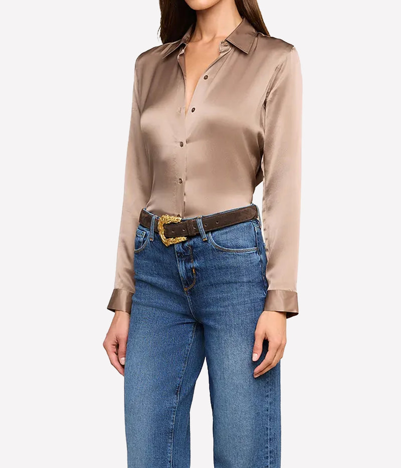 A woman wearing a long-sleeve silk button-up blouse in a taupe-brown colour, tucked into blue jeans with a dark brown belt featuring a gold buckle.