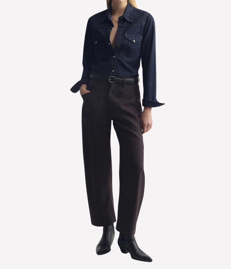 A model wearing a dark indigo denim shirt tucked into dark brown wide-leg trousers, paired with a black belt and black ankle boots, against a plain light gray background.
