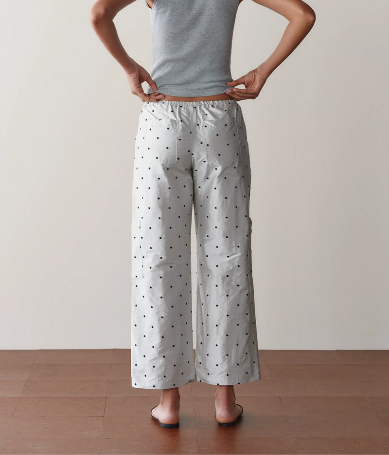 Back view of a model wearing the DONNI. The Silk Taffeta Cargo Pant in Silver Polka Dot with hands on hips and a grey tank top, showing the gathered waistband and wide cropped leg