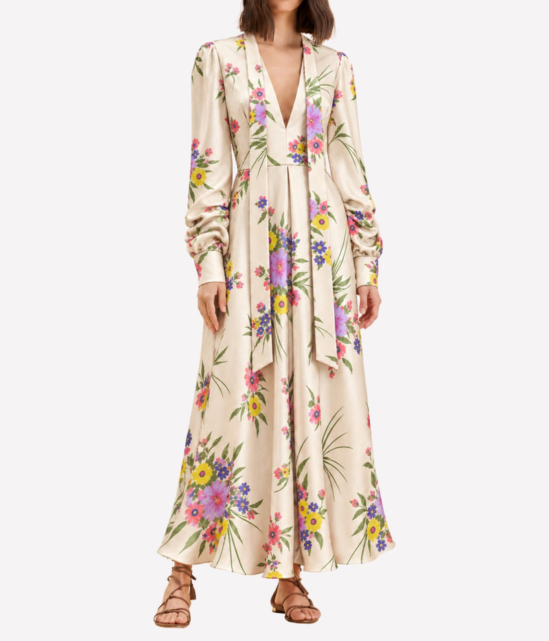 Front view of a model wearing the Raquel Diniz Valerie Dress in Pink Floral with the neck ties hanging loose and tan strappy sandals, showing the full maxi length