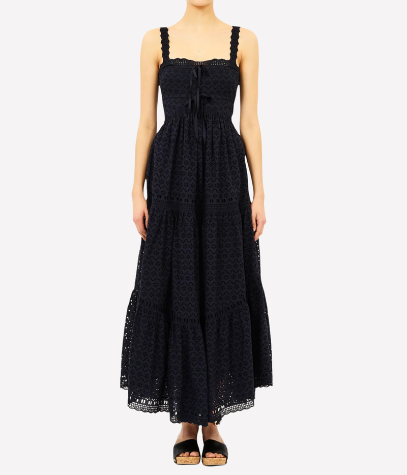 A woman modeling a black lace maxi dress with a tie-front bodice and tiered skirt, paired with black sandals, against a light gray studio background.