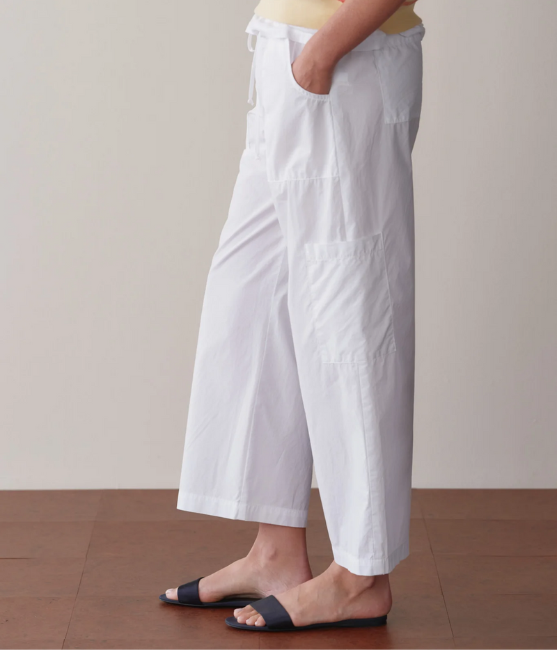 Side profile of a model wearing the DONNI. The Pop Drawstring Cargo Pant in Powder with a hand in the front pocket, showing the side cargo pocket, straight leg and cropped length with navy slide sandals