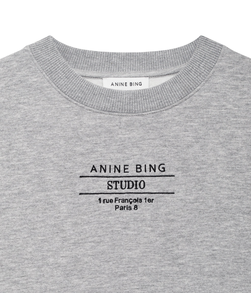 Close-up of a heather gray crewneck sweatshirt with black printed text reading 'ANINE BING STUDIO' and a small address below, on a white background.