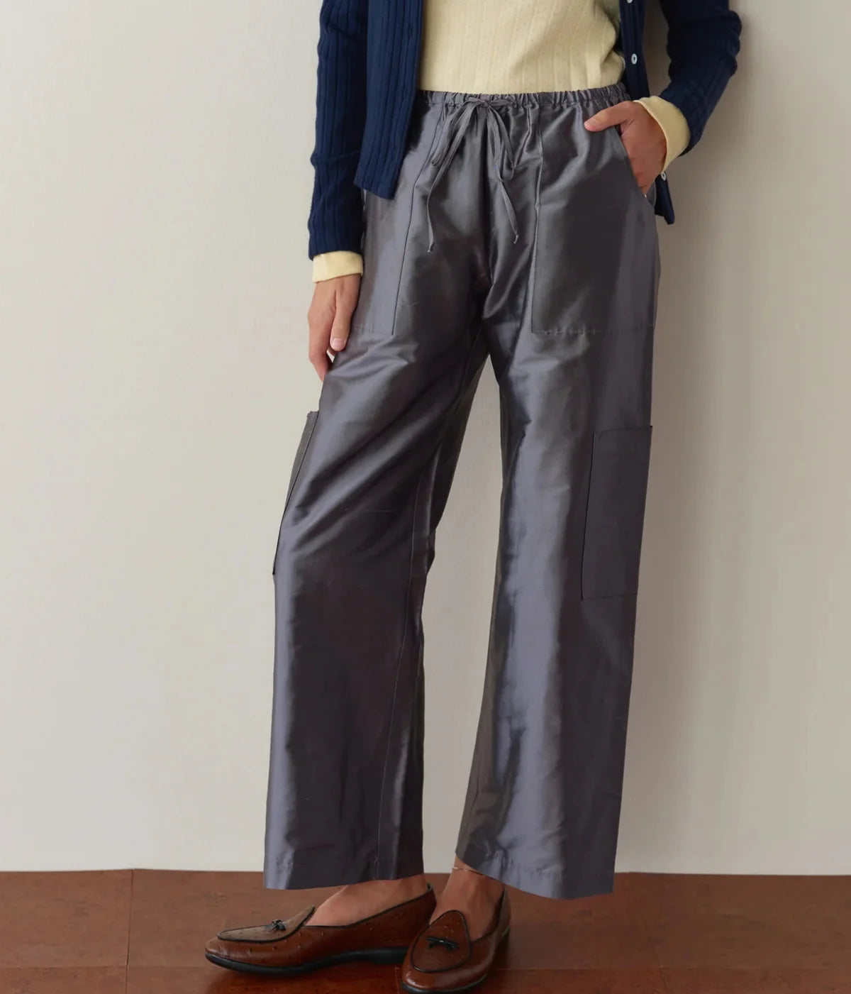 Front view of a model wearing the DONNI. The Silk Taffeta Cargo Pant in Pewter with one hand in the pocket, styled with a pale yellow ribbed top, open navy cable-knit cardigan and brown leather tassel loafers