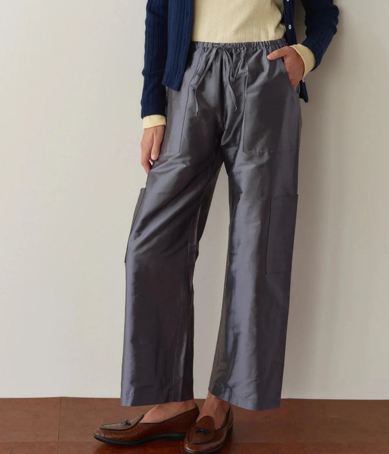 Front view of a model wearing the DONNI. The Silk Taffeta Cargo Pant in Pewter with one hand in the pocket, styled with a pale yellow ribbed top, open navy cable-knit cardigan and brown leather tassel loafers