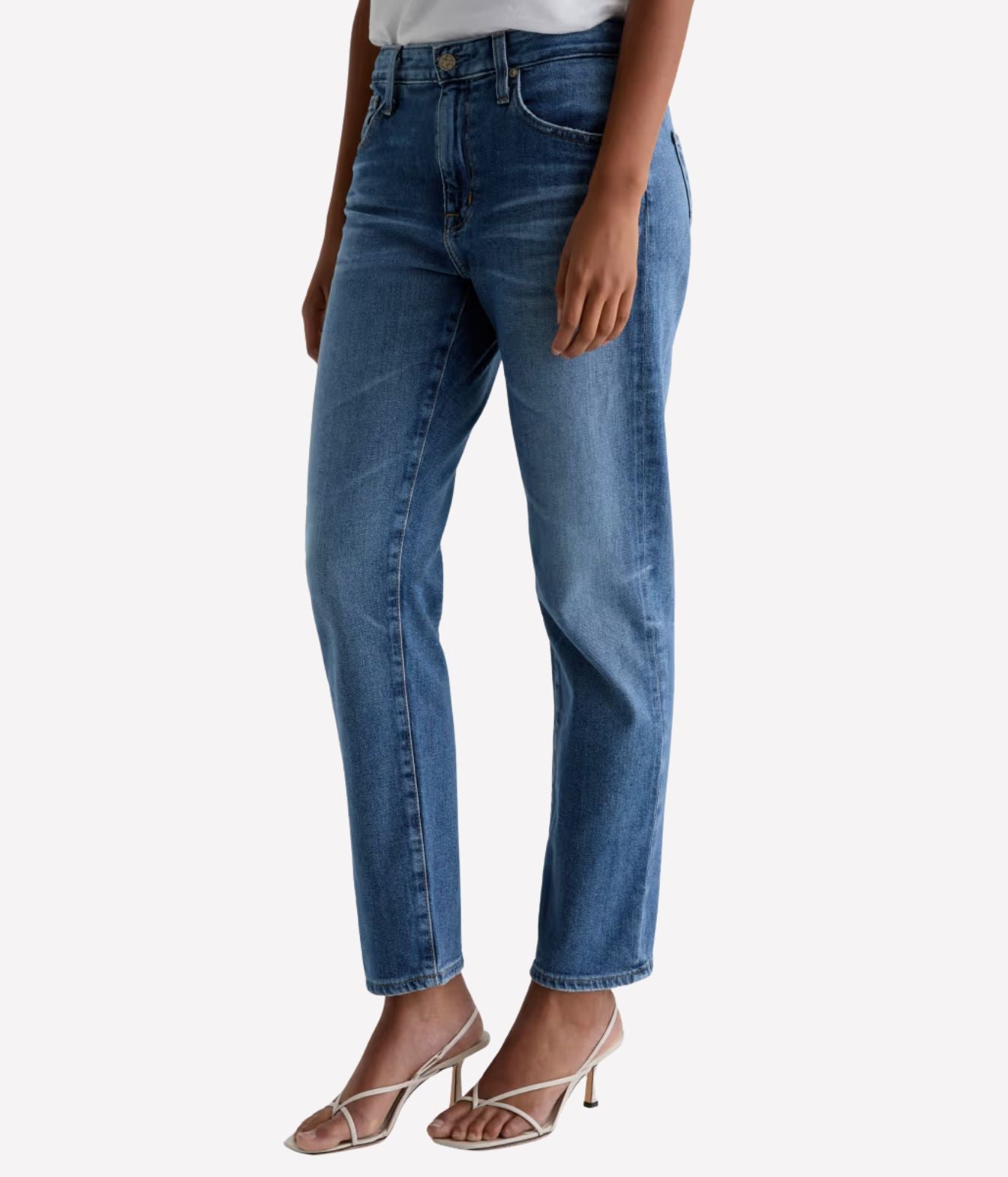 A model wearing a pair of medium-wash blue denim jeans with a straight-leg fit, paired with white strappy high-heeled sandals against a light gray background.
