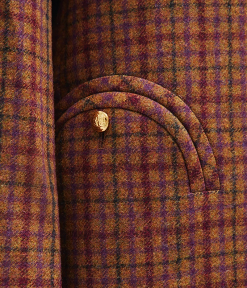 Close-up of a textured wool fabric with a purple and orange-brown plaid pattern, featuring a curved pocket with a single gold button