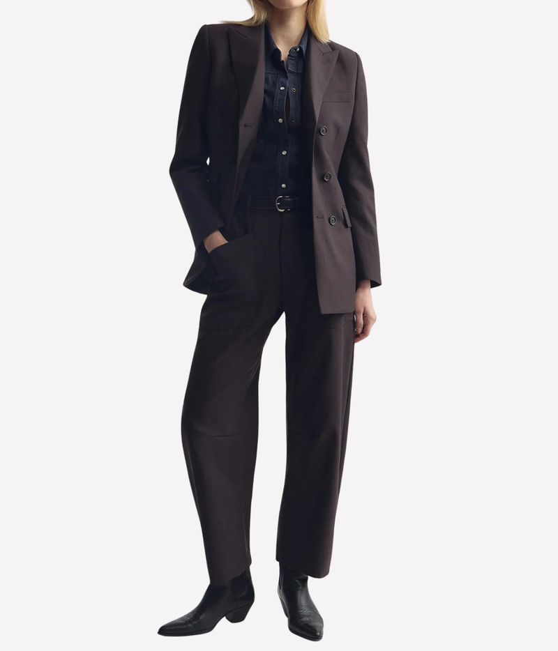 A model wearing a dark brown double-breasted blazer, matching trousers, a dark button-up shirt, and black ankle boots, shown against a light gray studio background.