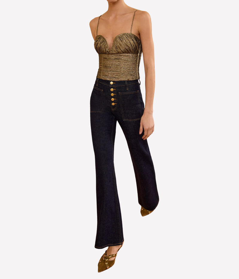 Full-length view of a model wearing the Ulla Johnson Vina Taffeta Cropped Bustier Top in Light Gold with dark indigo flared jeans with a gold button fly and pointed heels