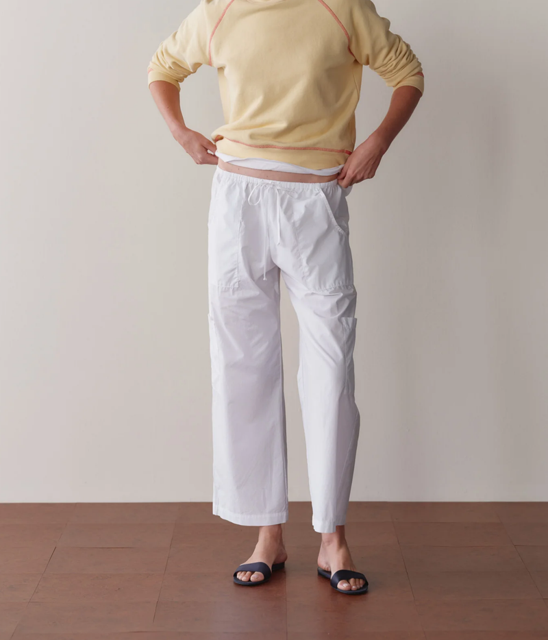Model wearing the DONNI. The Pop Drawstring Cargo Pant in Powder with hands on hips, showing the low-slung drawstring waist, styled with a pale yellow sweatshirt, white tee and navy slides