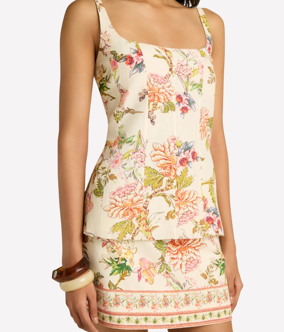 A model wearing a sleeveless peplum-style mini dress with a vibrant floral print on a cream background. The dress features pink, orange, and green flowers with a matching patterned border at the hem.