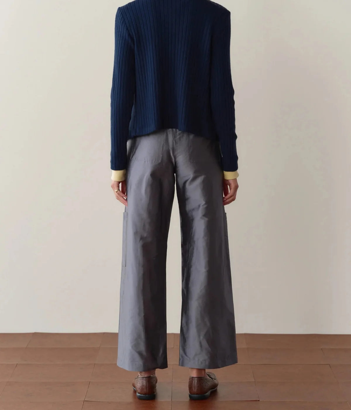 Back view of a model wearing the DONNI. The Silk Taffeta Cargo Pant in Pewter, showing the elasticated back waistband and straight leg, with a navy cardigan and brown loafers