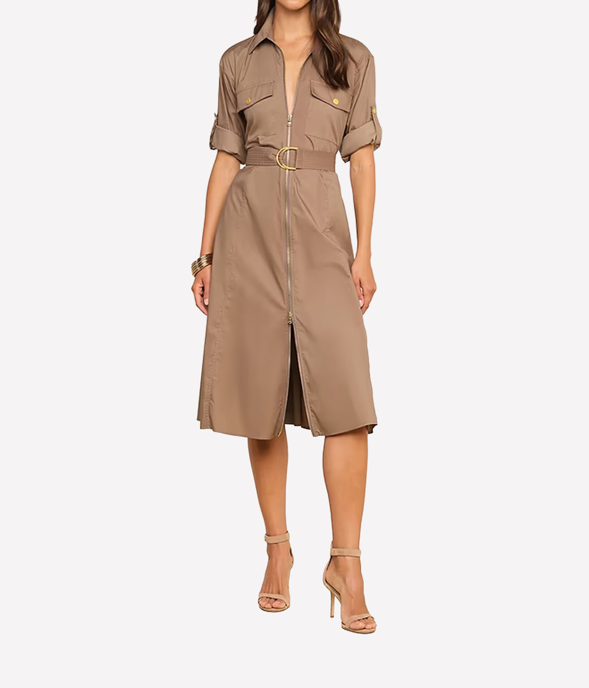 A woman modeling a taupe-brown utility shirt dress with a front zipper, chest pockets, rolled sleeves, and a matching belt with a gold buckle, paired with beige high-heeled sandals against a light grey background.