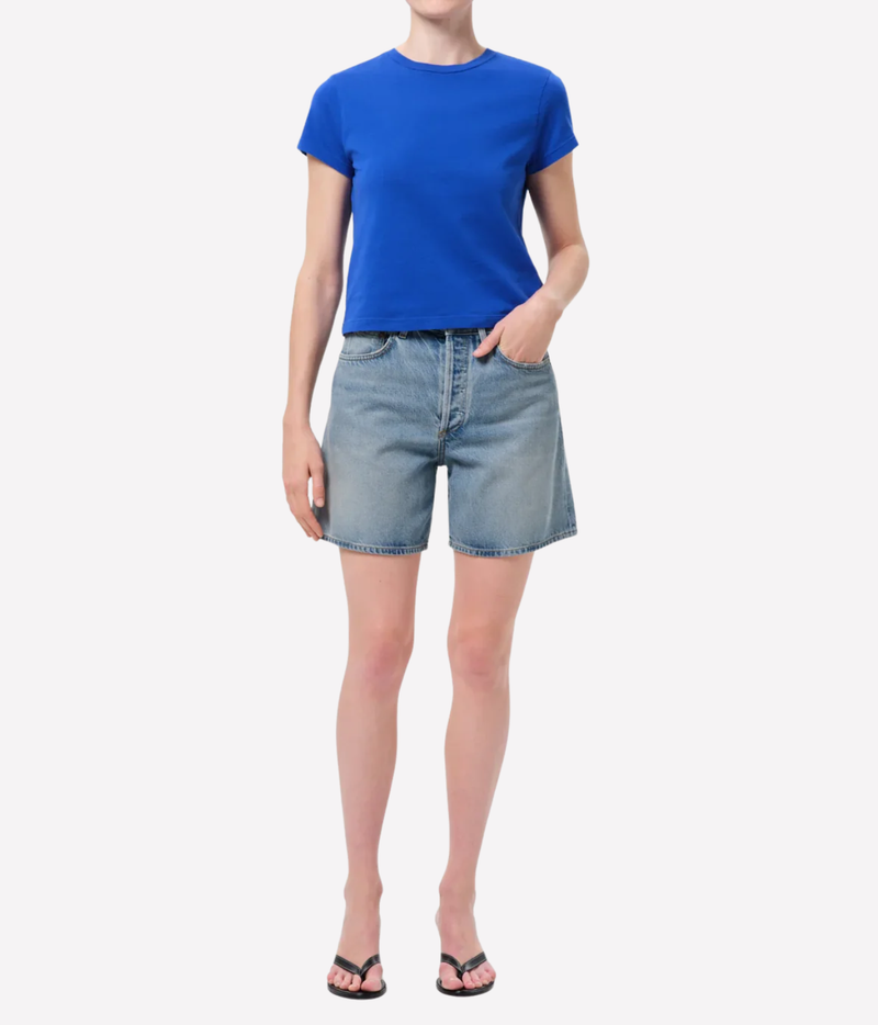 A model wearing a bright blue short-sleeve crewneck t-shirt and light-wash denim shorts, Agolde Dame Short in Reset