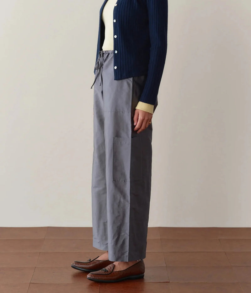 Side profile of a model wearing the DONNI. The Silk Taffeta Cargo Pant in Pewter, showing the relaxed leg and side cargo pocket, with a navy ribbed cardigan over a pale yellow top and brown tassel loafers