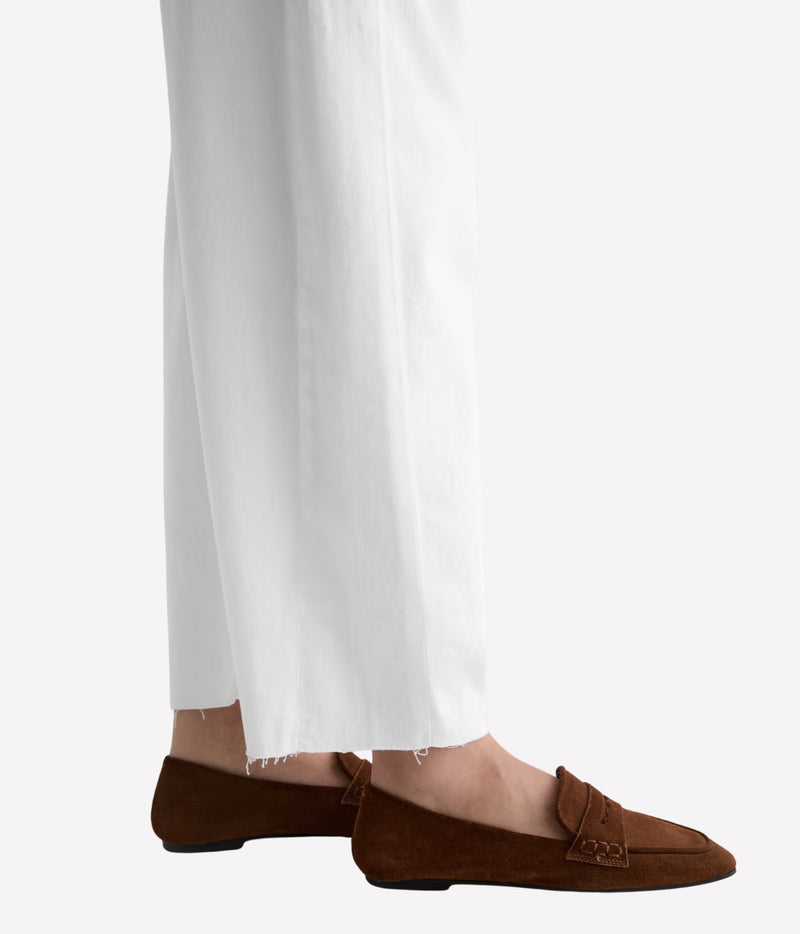 A close-up of a person's feet wearing brown suede penny loafers with white wide-leg trousers, shown against a light gray studio background.