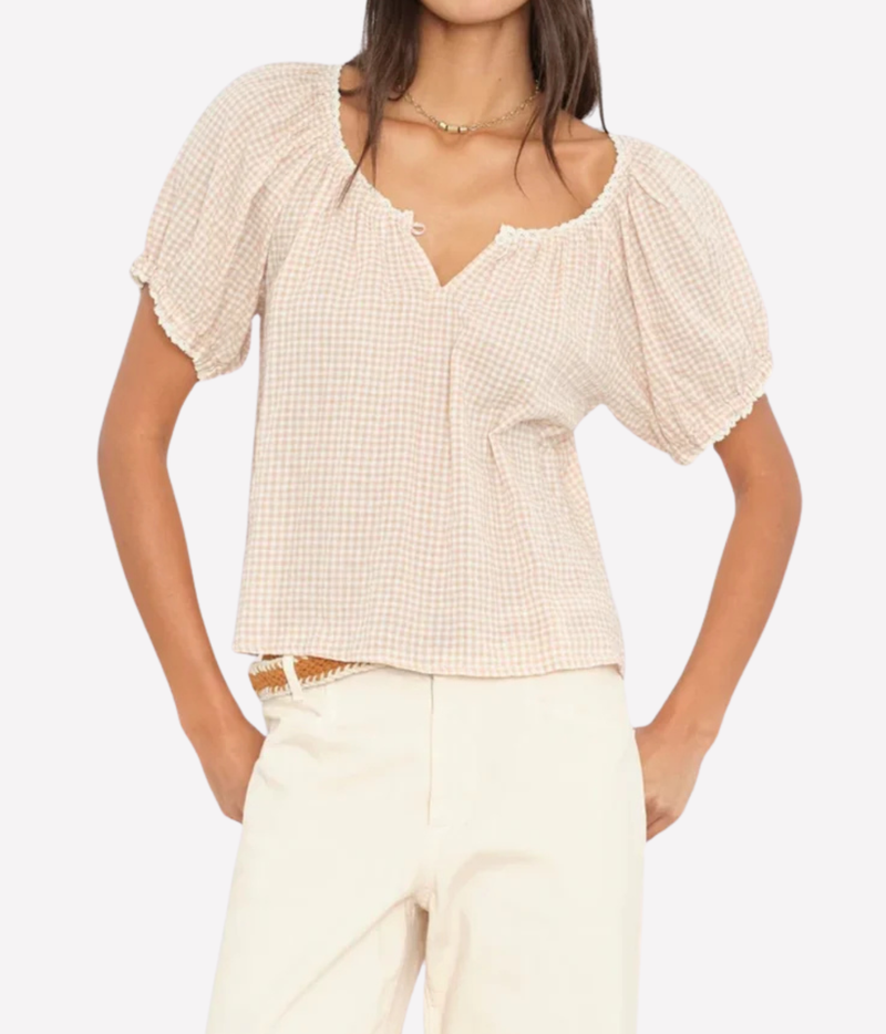 A woman wearing a cream and beige gingham short-sleeve blouse with puff sleeves and a split neckline, paired with cream pants, against a light gray studio background.