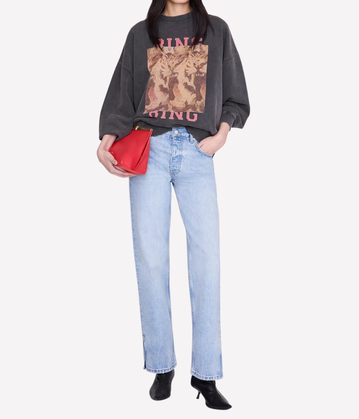 A model wearing a washed black graphic sweatshirt, light-wash blue straight-leg jeans, and black ankle boots, holding a red clutch bag against a plain light gray background.