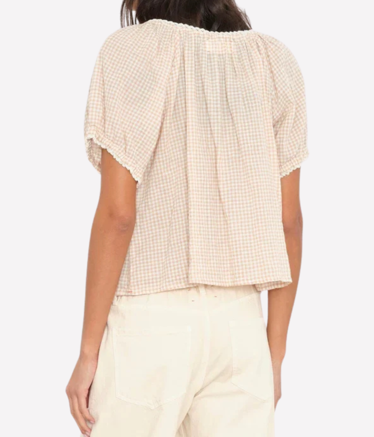 A woman shown from the back wearing a short-sleeve cream and beige gingham blouse with a ruffled neckline and pleated back, paired with light cream pants, against a plain light gray background.