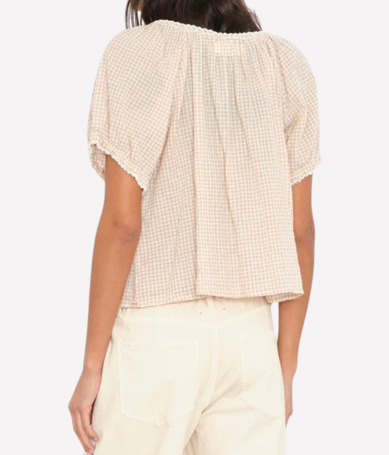 A woman shown from the back wearing a short-sleeve cream and beige gingham blouse with a ruffled neckline and pleated back, paired with light cream pants, against a plain light gray background.