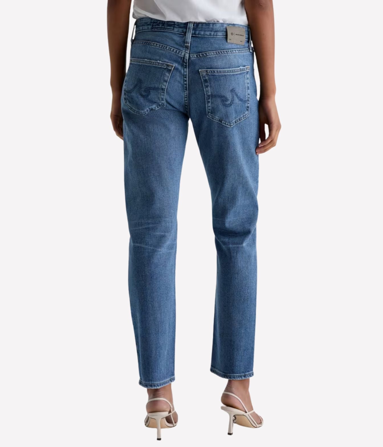 Rear view of a model wearing medium-wash blue denim jeans with a straight-to-slim leg, paired with a white top and beige high-heeled sandals, against a light gray background.