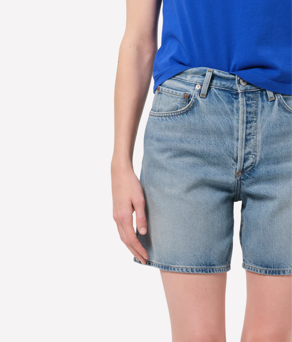 A close-up of a person wearing light-wash blue denim shorts and a bright blue top, Agolde Dame Short in Reset