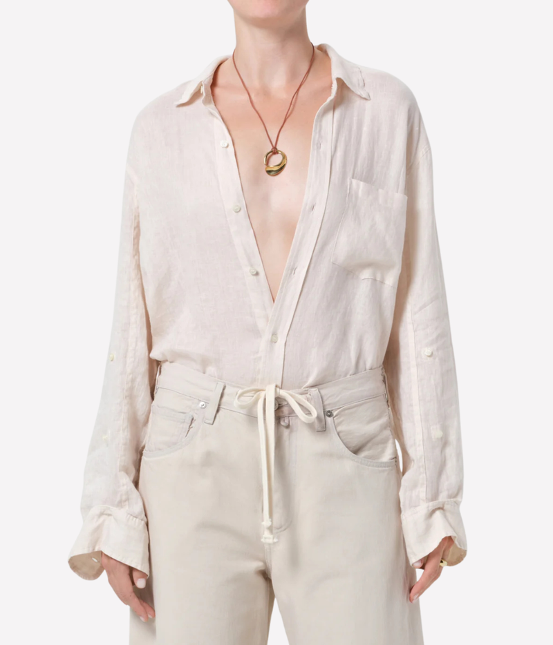 A model wearing a light beige linen button-down shirt with a chest pocket and matching beige drawstring trousers, styled with a gold pendant necklace against a plain white background.