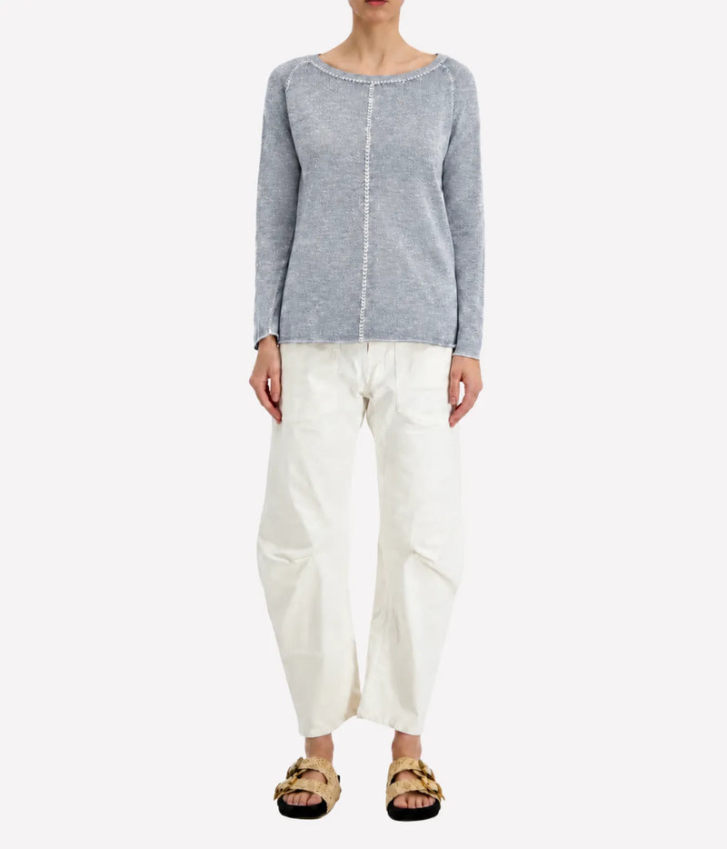 Front view of a model wearing the Avant Toi Maglia Pullover In Dusty Blue untucked over cream barrel-leg trousers with dark sandals, showing the raglan sleeves and relaxed fit