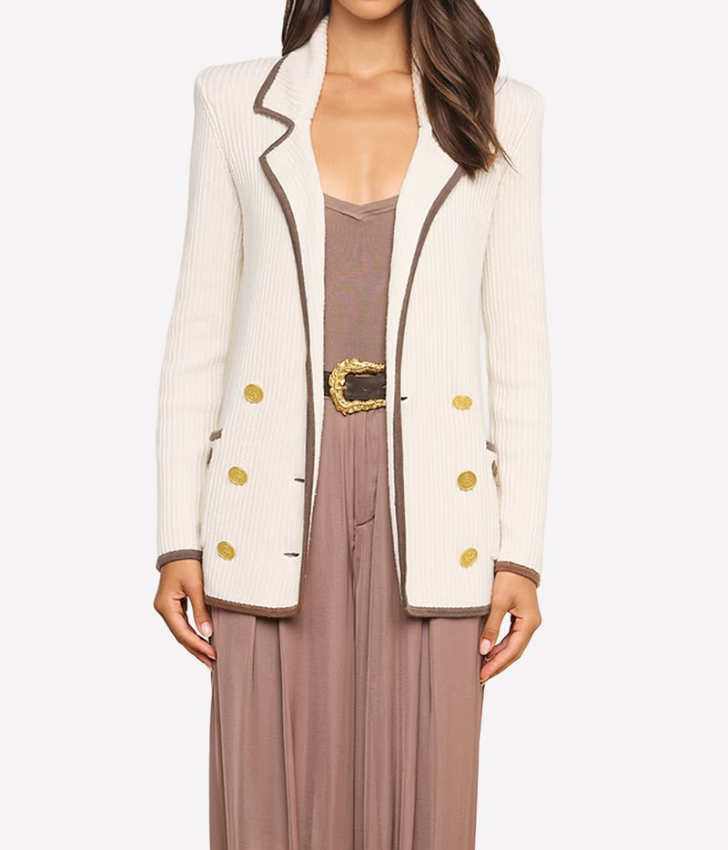 A woman wearing a cream knit blazer with brown trim and gold buttons over a brown beige top.
