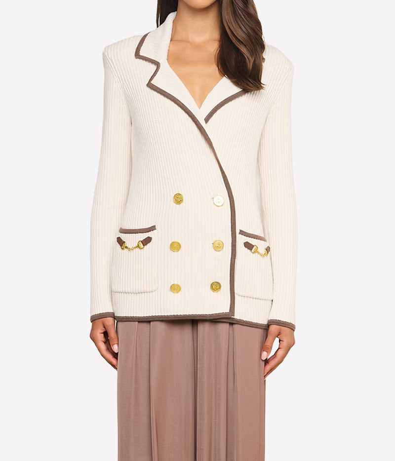 A woman wearing a cream ribbed knit blazer with brown trim and gold buttons, paired with taupe wide-leg pants.