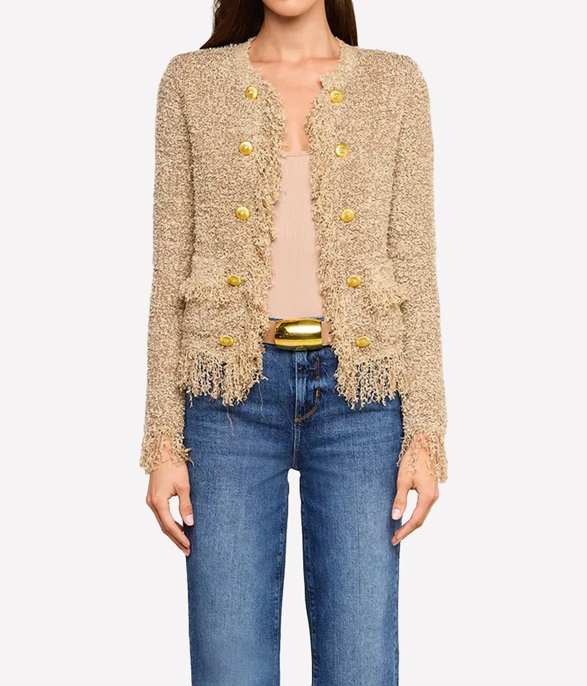 A woman wearing the Mireille Fringe Cardigan with textured beige fabric with metallic threads and gold buttons, paired with a light-coloured top and blue jeans.