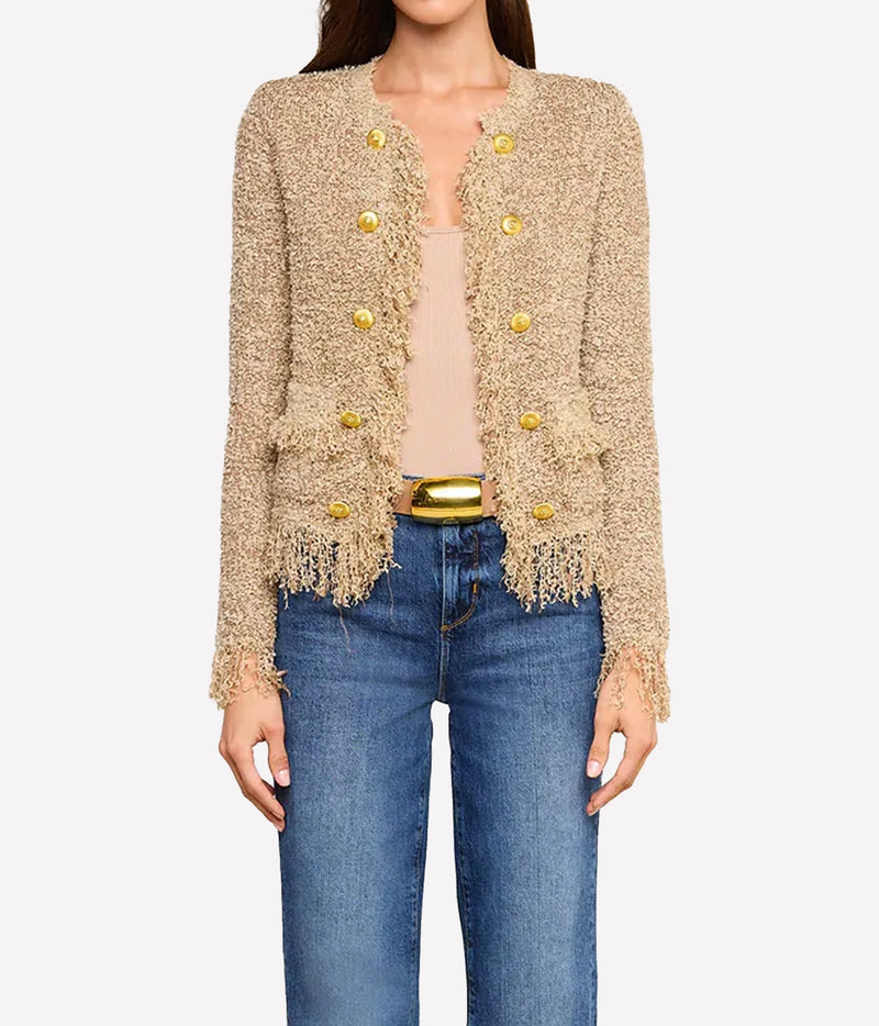 A woman wearing the Mireille Fringe Cardigan with textured beige fabric with metallic threads and gold buttons, paired with a light-coloured top and blue jeans.
