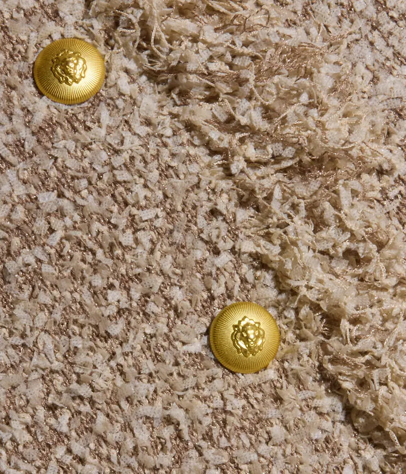 Close-up of the Mireille Fringe Cardigan’s textured beige confetti knit fabric, featuring two shiny gold lion’s head buttons and diagonal sections of smooth and fringed detailing.