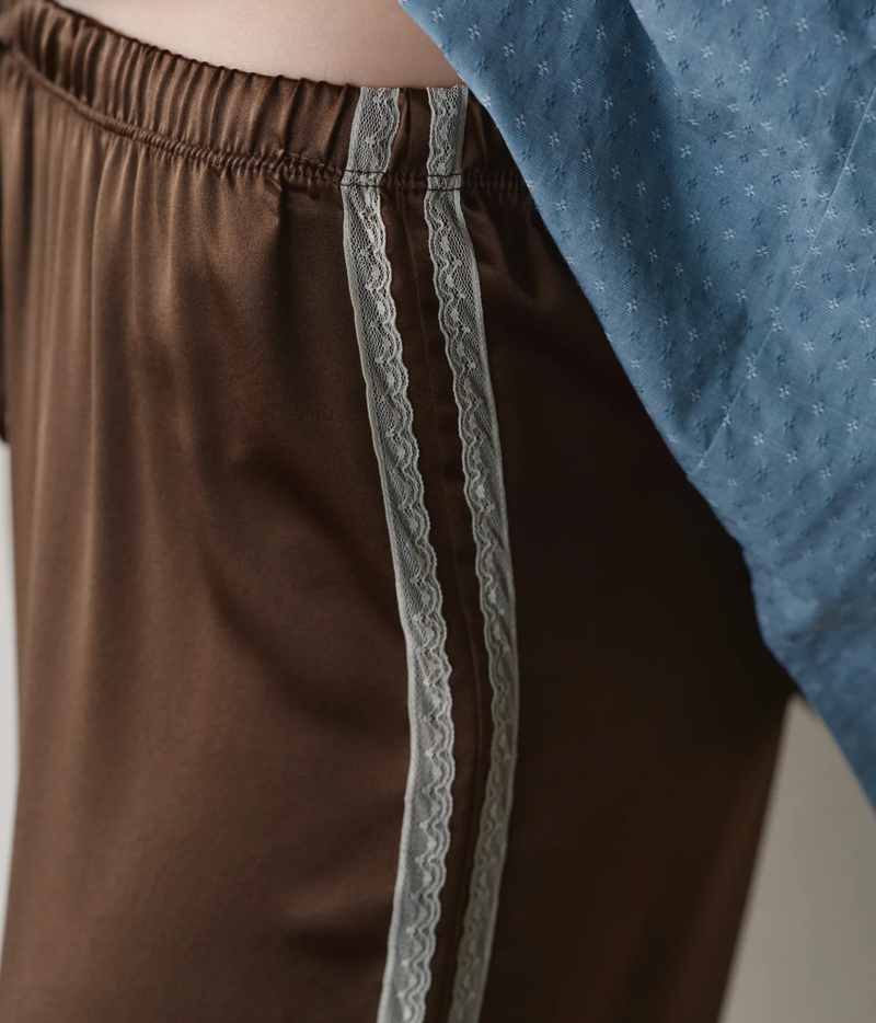 Close-up of the DONNI. The Silk Lace Stripe Crop Pant in Espresso showing the gathered elastic waistband and twin ivory lace stripes against the silk, beside the hem of a blue printed shirt