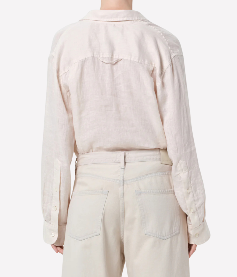 Back view of a person wearing a light cream linen shirt and matching cream trousers against a plain light gray studio background.