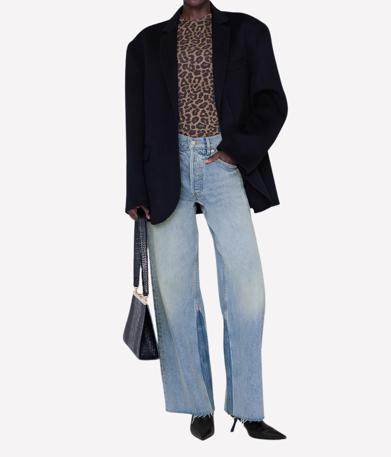 A model wearing an oversized black blazer, light-wash wide-leg denim jeans, a brown and black leopard print top, and black pointed-toe heels, holding a black handbag against a plain light gray background.
