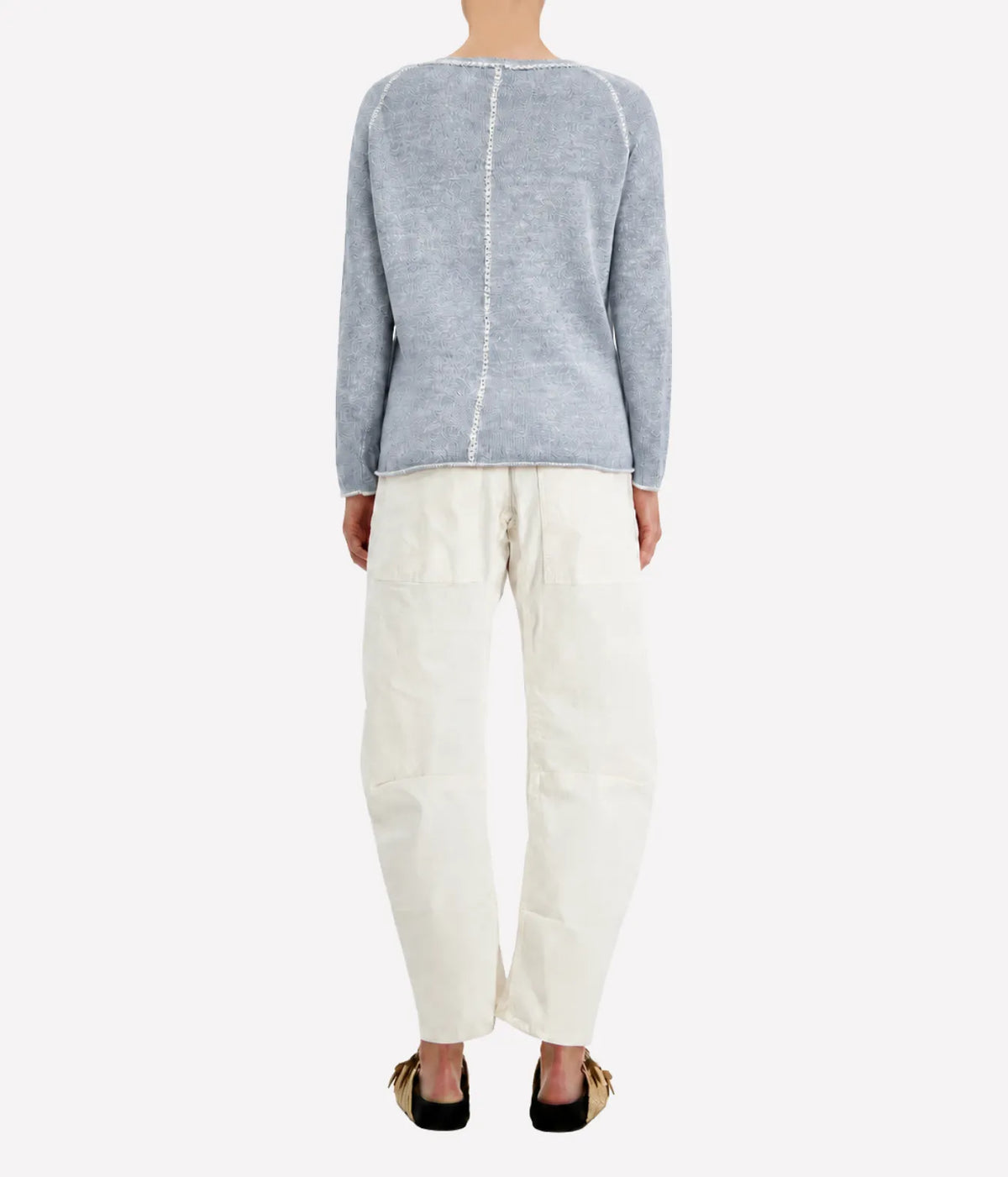 Back view of a model wearing the Avant Toi Maglia Pullover In Dusty Blue with cream barrel-leg trousers and dark sandals, showing the contrast centre-back seam