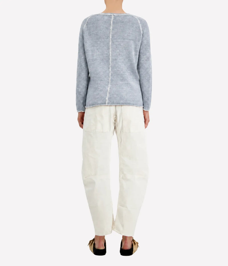 Back view of a model wearing the Avant Toi Maglia Pullover In Dusty Blue with cream barrel-leg trousers and dark sandals, showing the contrast centre-back seam